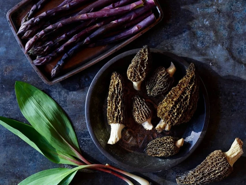 Overhead shot of morel mushrooms on a black plate, purple asparagus on a tray, and wild garlic leaves on a dark, textured surface.