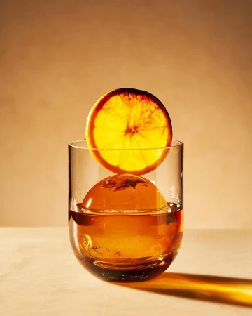 Still life of a cocktail glass filled with ice and amber liquor, topped with a vibrant orange slice against a warm backdrop.