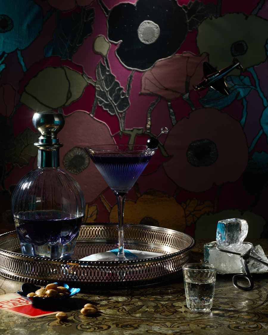 Still life photo of a cocktail in a martini glass and a decanter with a purple liquid on a silver tray with almonds and ice.