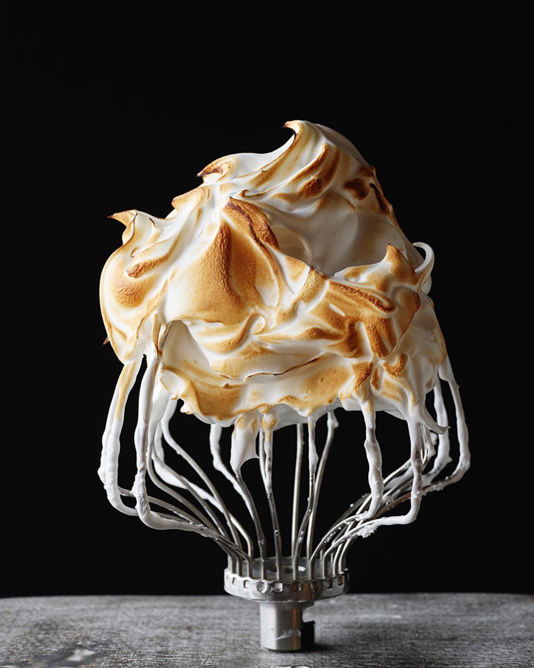 Close-up of a toasted meringue dessert piped onto a metal whisk, set against a stark black background for a minimalist food still life.