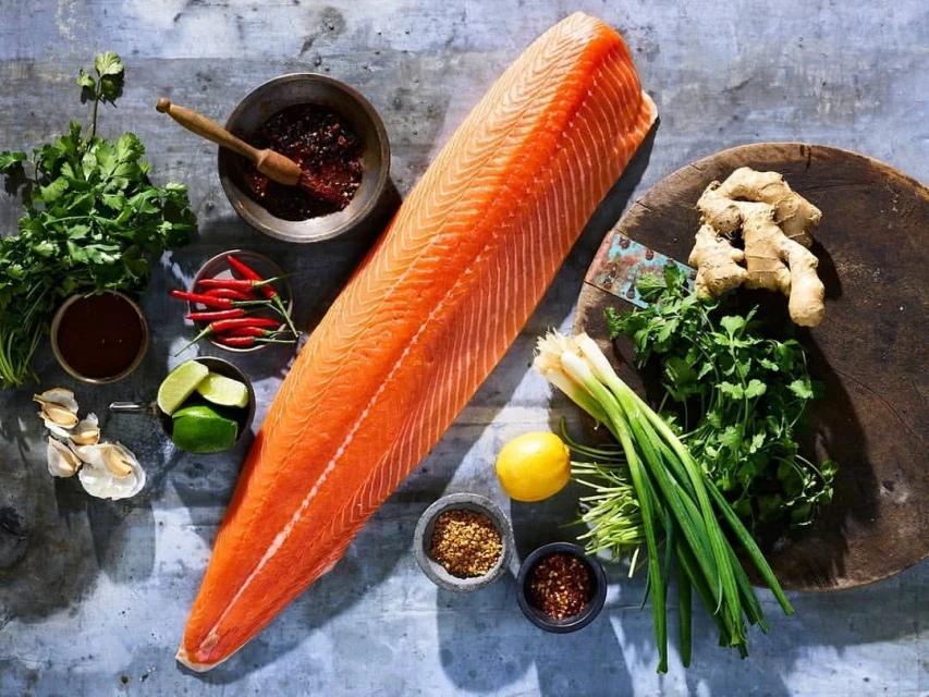 Overhead shot of fresh salmon fillet surrounded by ingredients like ginger, chili peppers, garlic, cilantro, and spices on grey surface.