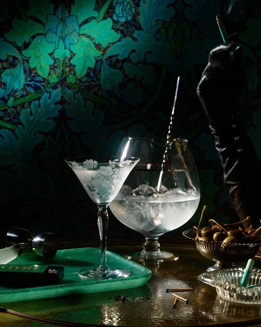Two crystal cocktail glasses, one filled with ice and a black gloved hand holding a cigarette against a dark floral wallpaper.