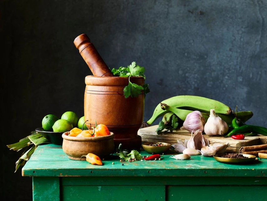 Wooden mortar and pestle with plantains, limes, peppers, and spices on a green table against a dark background, creating a food still life.