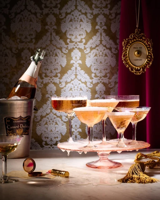 Celebratory image featuring champagne glasses, an ice bucket with a champagne bottle, and vintage-style props against a damask wallpaper backdrop.