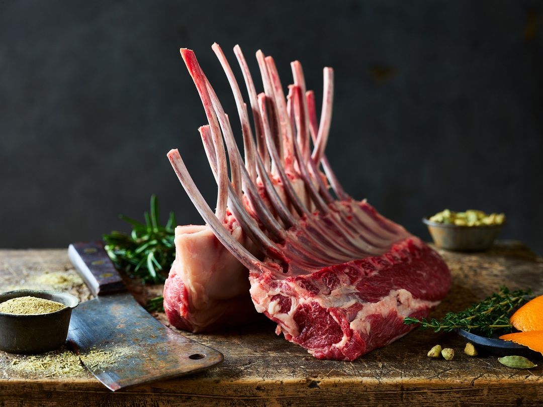 Close-up of a raw rack of lamb, herbs, and knife on a wooden cutting board against a dark background, food photography for a butcher shop.