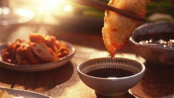 Close-up shot of a piece of fried food being dipped into a bowl of dark sauce with food in the blurred background.