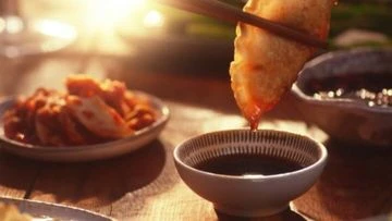 Close-up shot of a piece of fried food being dipped into a bowl of dark sauce with food in the blurred background.