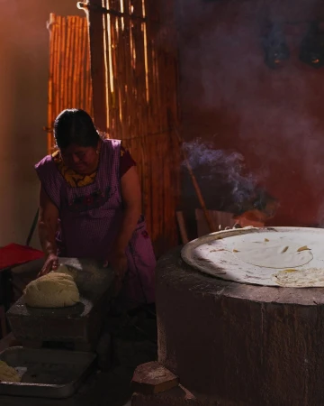 Woman prepares food in a rustic kitchen. She's pressing dough on a stone, with a stove and cooking surface emitting steam in the background.