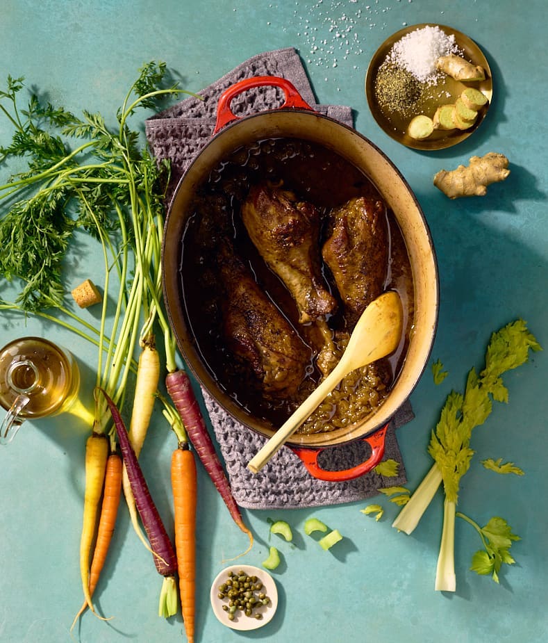 Overhead shot of a stew in a cast iron pot surrounded by carrots, celery, ginger, and seasonings on a blue surface, studio lighting.
