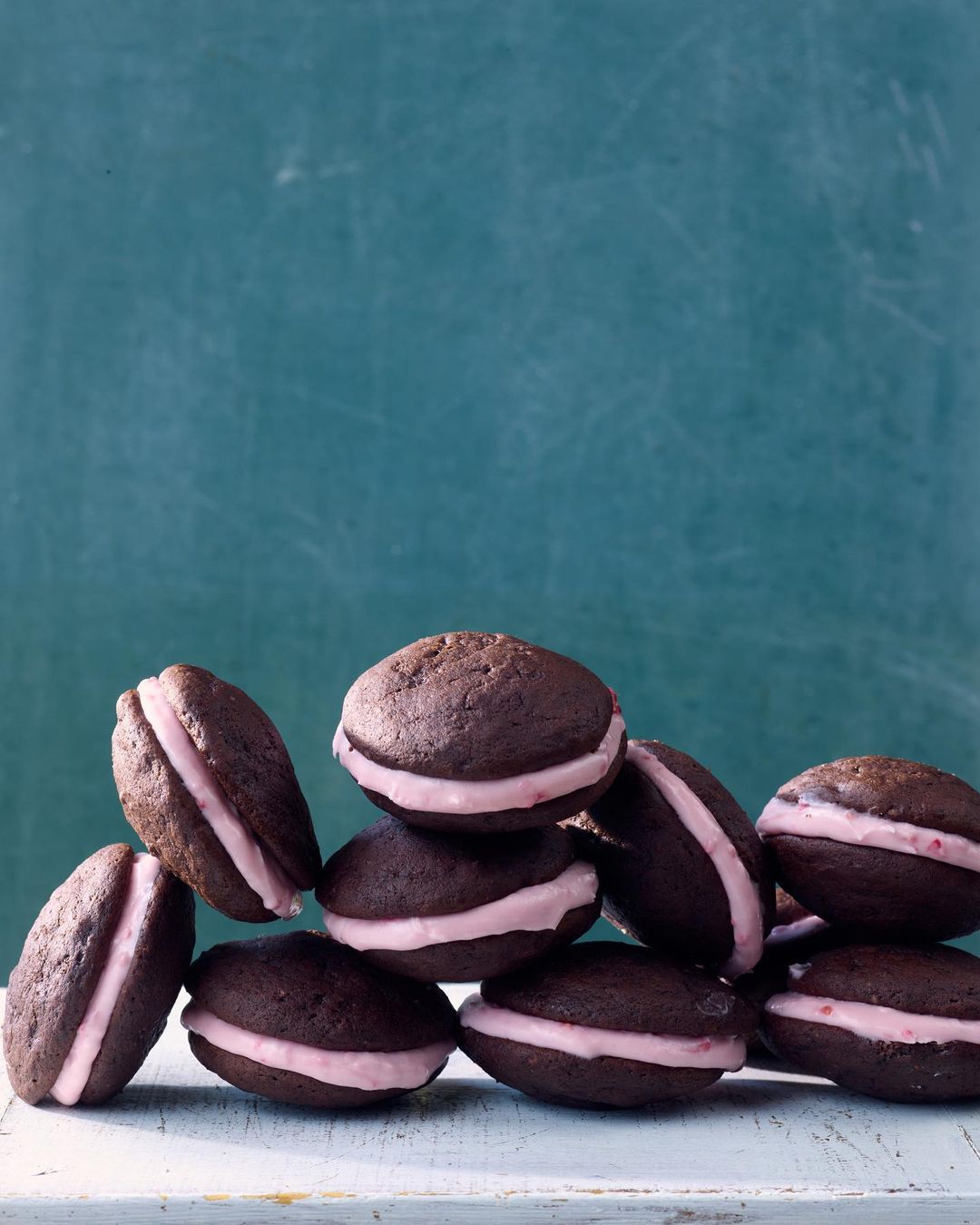 Pile of chocolate whoopie pies with pink filling stacked on a distressed white wooden surface against a turquoise backdrop.