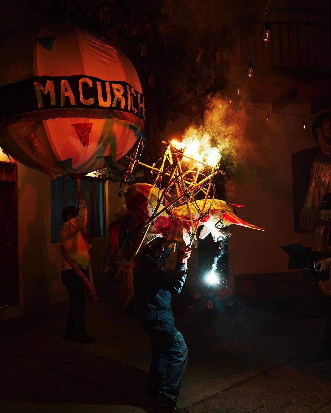 Procession with a man carrying a burning sculpture and a person holding a globe-shaped paper lantern in a dark outdoor setting.