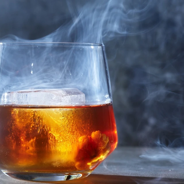 A tumbler glass holds a beverage with an ice cube, surrounded by smoke against a gray background in a studio shot for advertising.