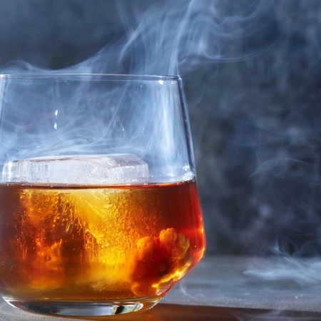 A tumbler glass holds a beverage with an ice cube, surrounded by smoke against a gray background in a studio shot for advertising.