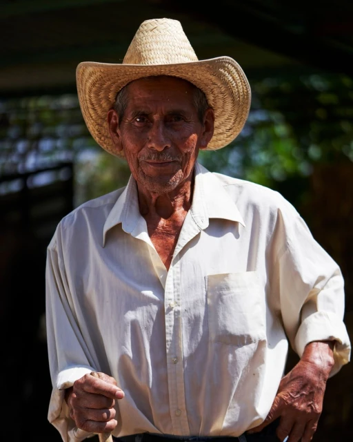 Portrait of an elderly man with tan skin wearing a straw hat and white button-down shirt, standing outdoors in natural light.