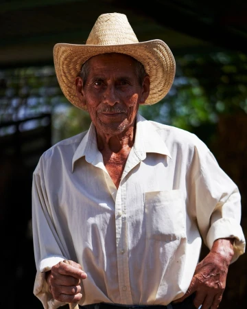 Portrait of an elderly man with tan skin wearing a straw hat and white button-down shirt, standing outdoors in natural light.