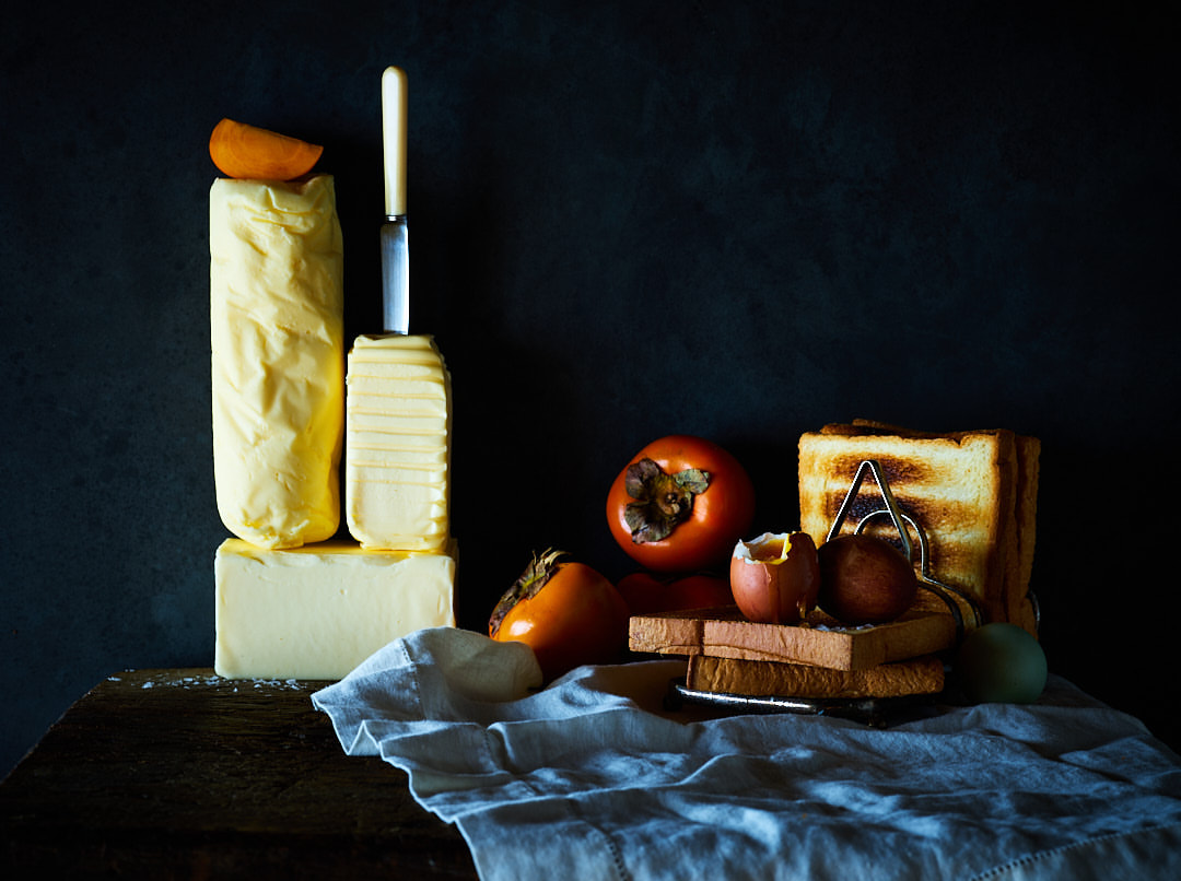 Still life photograph of butter blocks and sliced, toasted bread alongside persimmons and boiled eggs on a wooden surface.