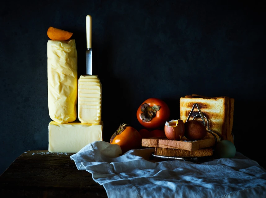 Still life photograph of butter blocks and sliced, toasted bread alongside persimmons and boiled eggs on a wooden surface.