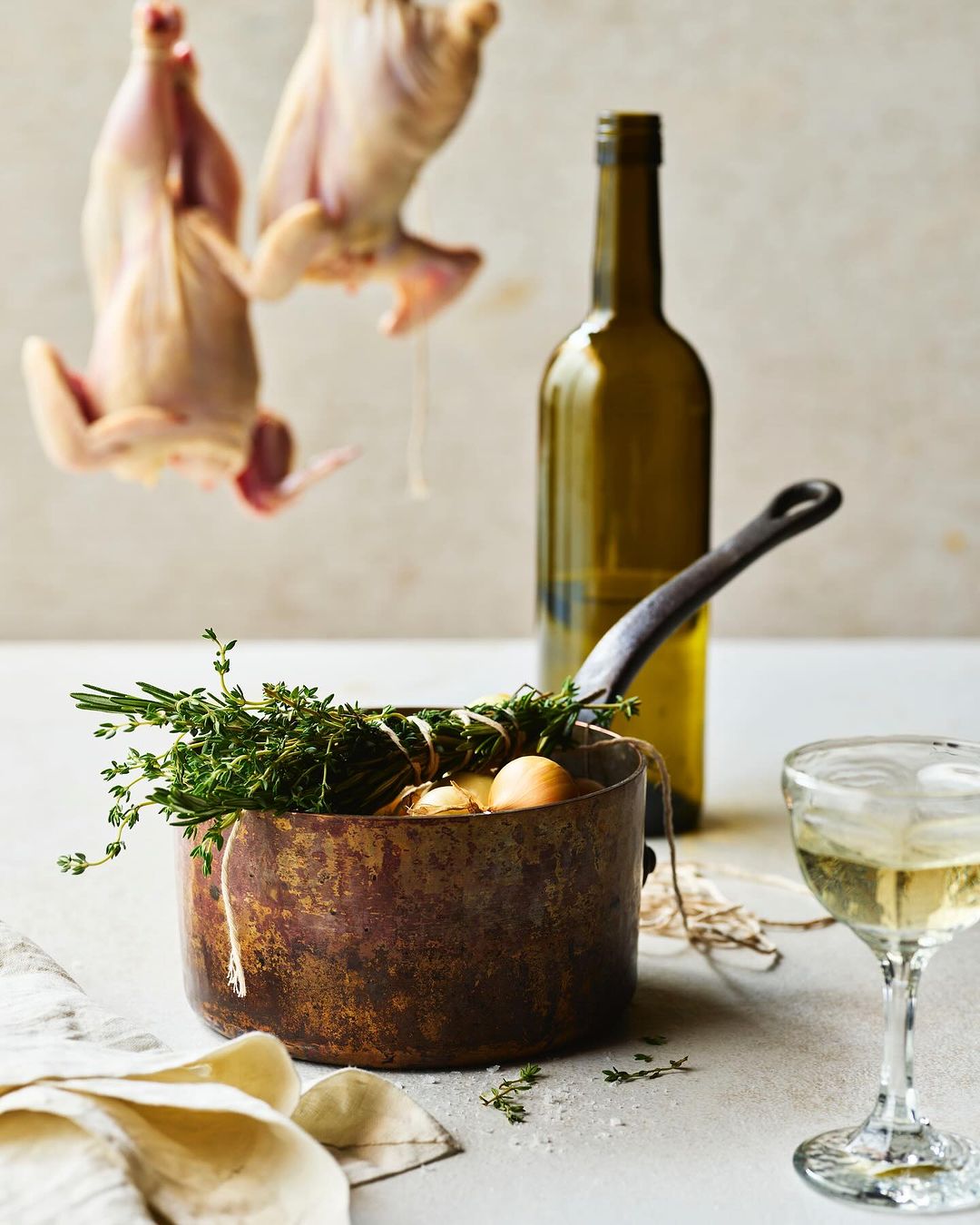 Rustic food still life features cooked game birds hanging over a copper pot of onions, a wine bottle, and a glass of white wine.