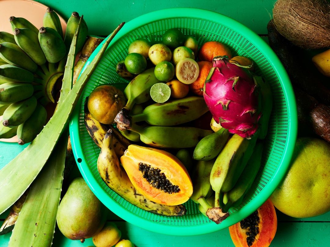 Overhead shot of a vibrant mix of tropical fruits like bananas, papaya, dragon fruit, and limes arranged in a green plastic basket.