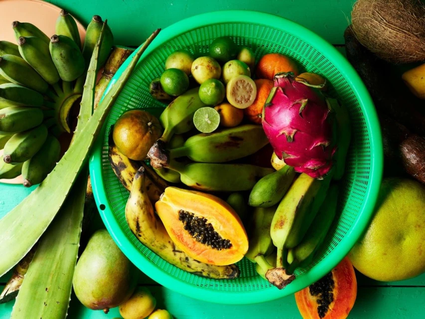 Overhead shot of a vibrant mix of tropical fruits like bananas, papaya, dragon fruit, and limes arranged in a green plastic basket.