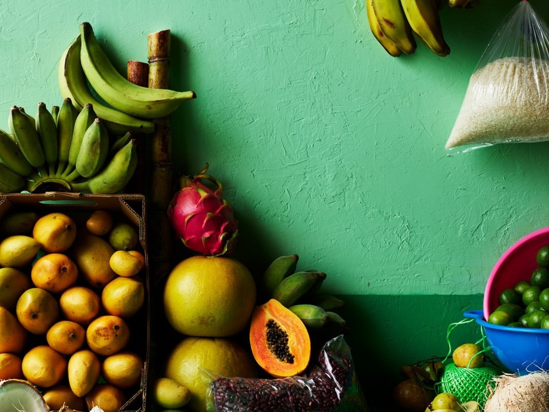 Still life shot of tropical fruits including bananas, mangos, papaya, dragon fruit, limes, and coconuts displayed against a green wall.
