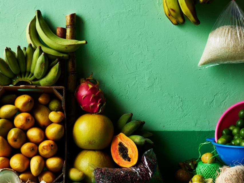 Still life shot of tropical fruits including bananas, mangos, papaya, dragon fruit, limes, and coconuts displayed against a green wall.