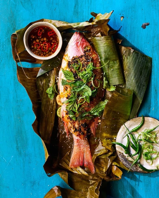 Overhead shot of red snapper with herbs and chillies on banana leaves, accompanied by dipping sauce on a vibrant turquoise background.