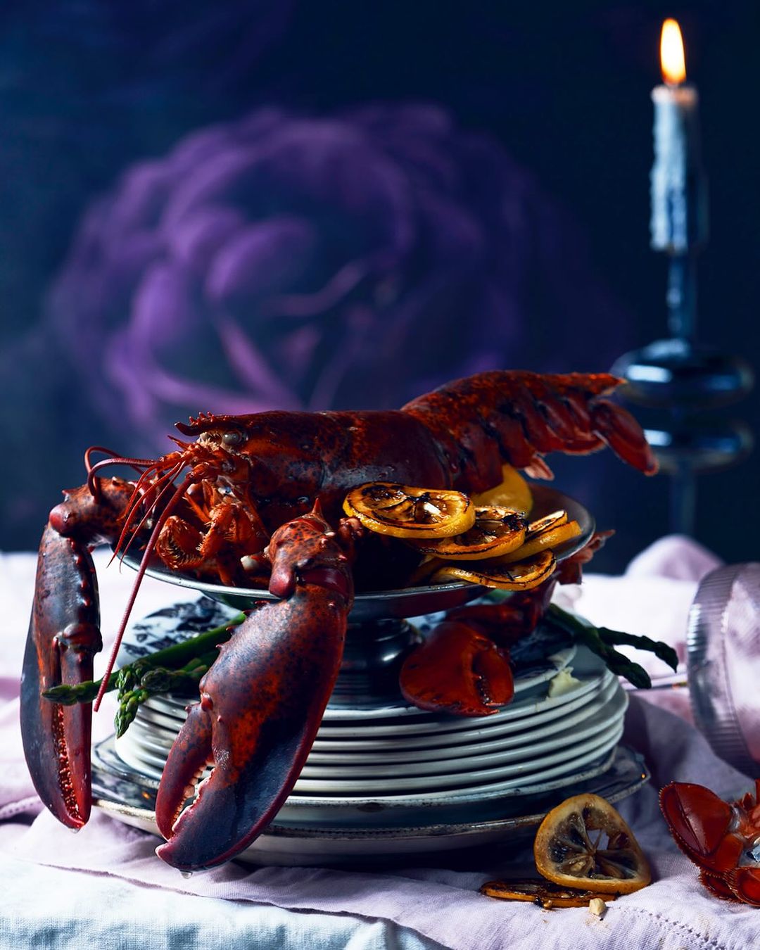 Dramatic food still life featuring a cooked lobster with grilled lemon slices on a stack of plates next to a burning candle.