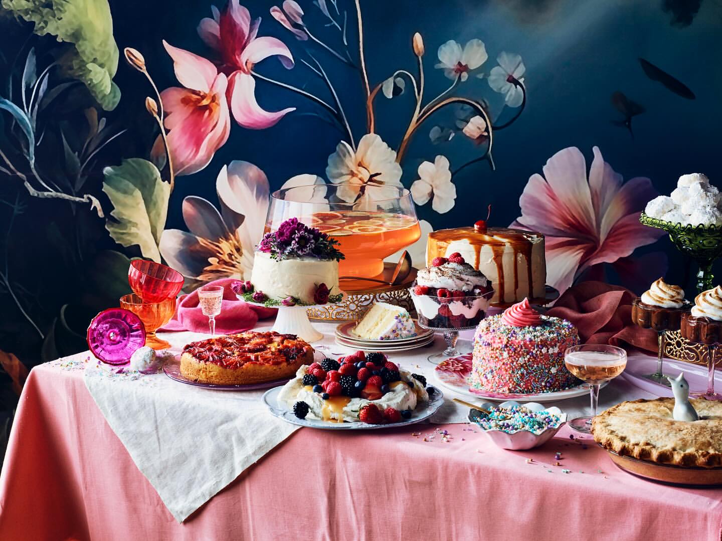 Elaborate dessert spread featuring assorted cakes, pies, drinks, and glassware on a pink tablecloth against a floral backdrop.