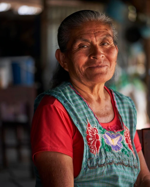 Portrait of a smiling older woman wearing a blue and green embroidered apron and a red t-shirt in an indoor setting with warm lighting.