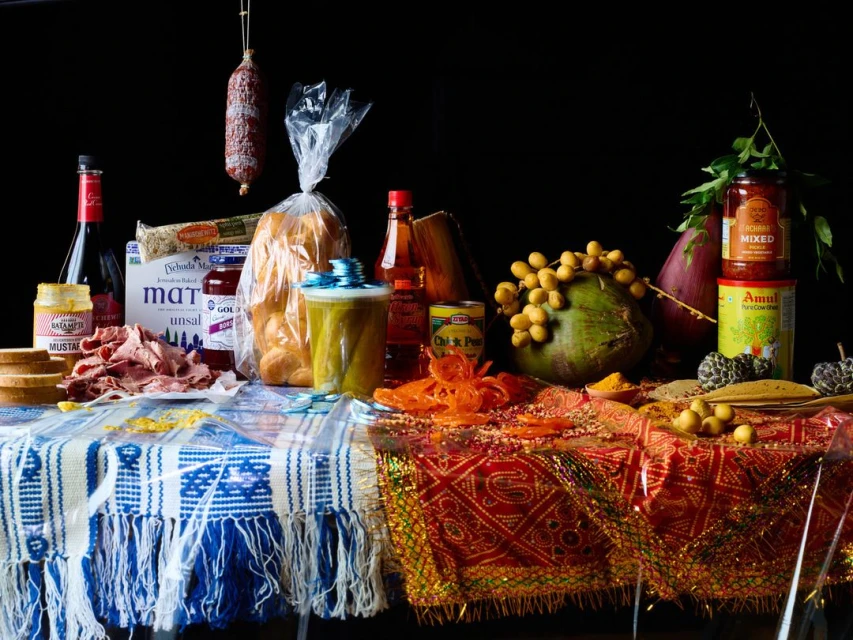 Food still life featuring a table laden with breads, meats, fruits, and packaged goods, illuminated by studio lighting against a dark backdrop.