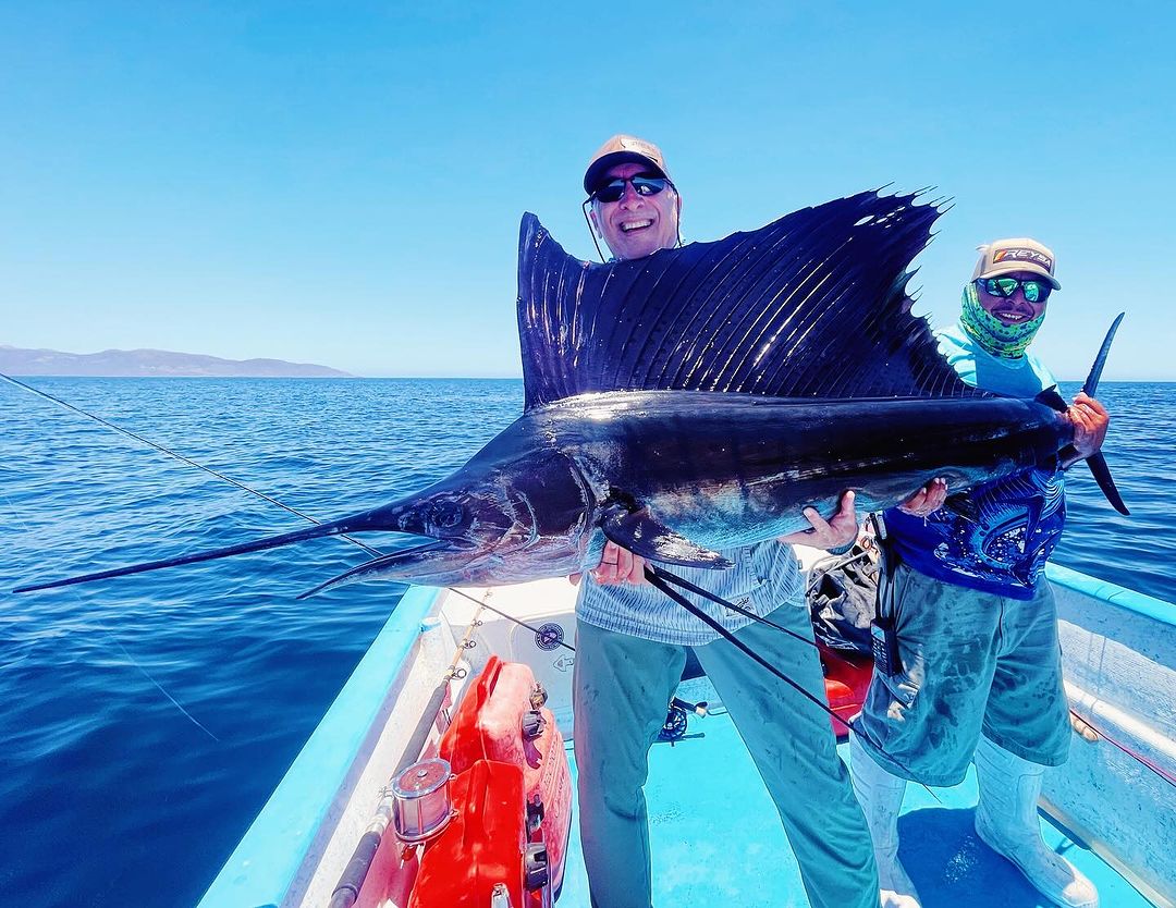 Two adult males on a boat holding a large sailfish against a backdrop of blue ocean and clear sky, showcasing a successful fishing trip.