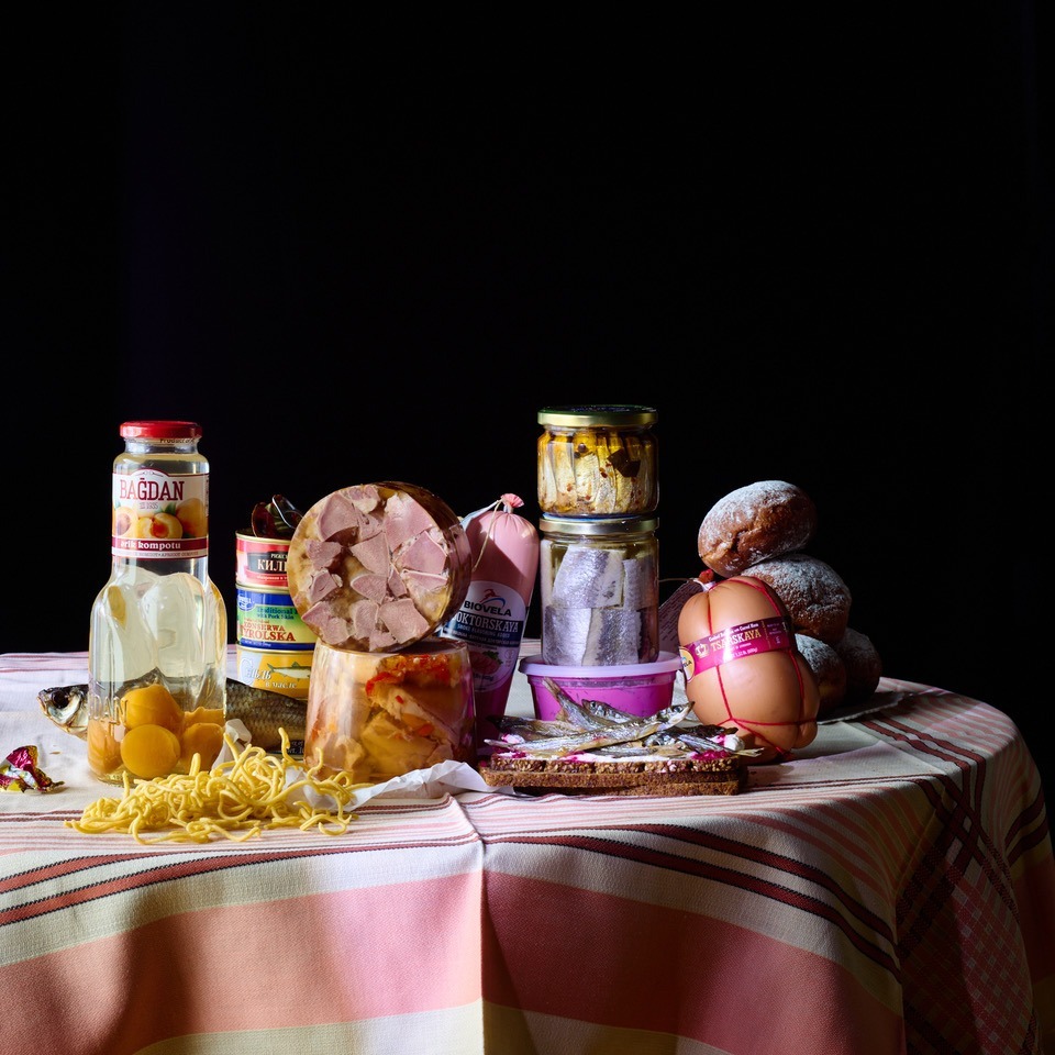 Still life of assorted preserved food products in jars, cans, and wrapped on a table against a dark background.