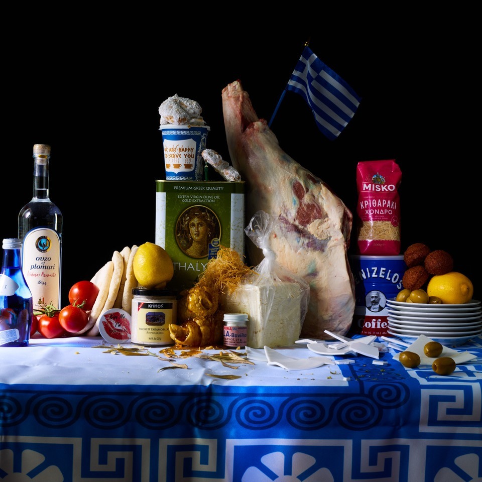Still life of various Greek food items including a leg of lamb, cheese, lemons, olive oil, and Greek flag against a black background.