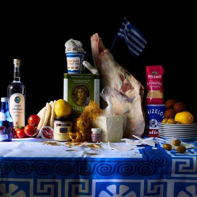 Still life of various Greek food items including a leg of lamb, cheese, lemons, olive oil, and Greek flag against a black background.