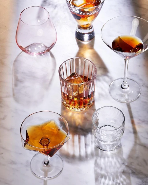 Six various clear drinking glasses, including martini, lowball, and wine glasses, some filled with cocktails, displayed on marble.