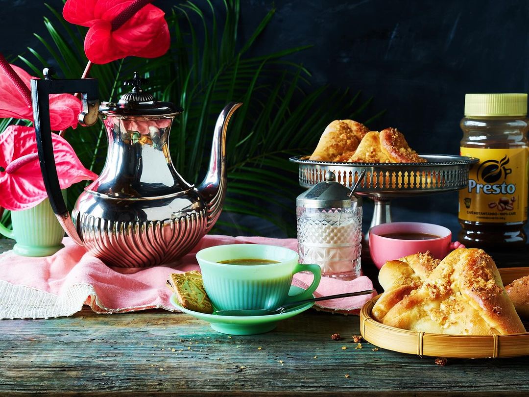 Silver tea set with pastries, coffee jar, and red anthurium flowers arranged on a rustic wooden table against a dark background.