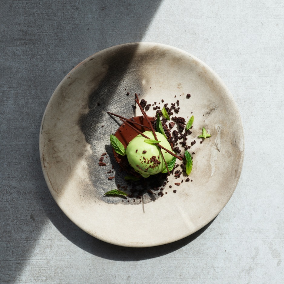 Overhead shot of a dessert featuring a light green scoop, chocolate sauce, mint leaves, and chocolate crumbles on a grey-toned plate.