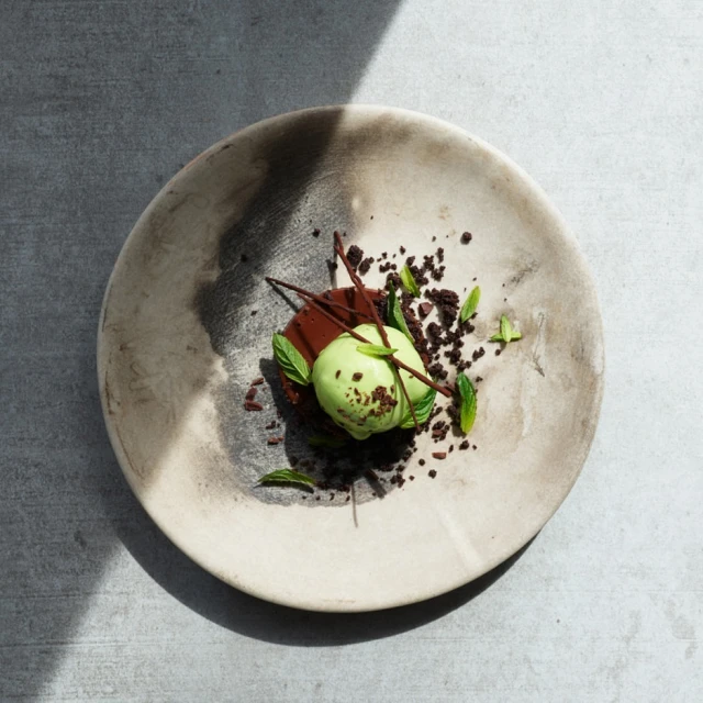 Overhead shot of a dessert featuring a light green scoop, chocolate sauce, mint leaves, and chocolate crumbles on a grey-toned plate.
