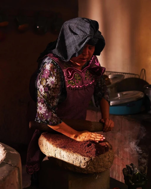 A woman in traditional clothing grinds spices on a stone. The indoor kitchen scene features warm, muted tones and a focus on cultural food preparation.