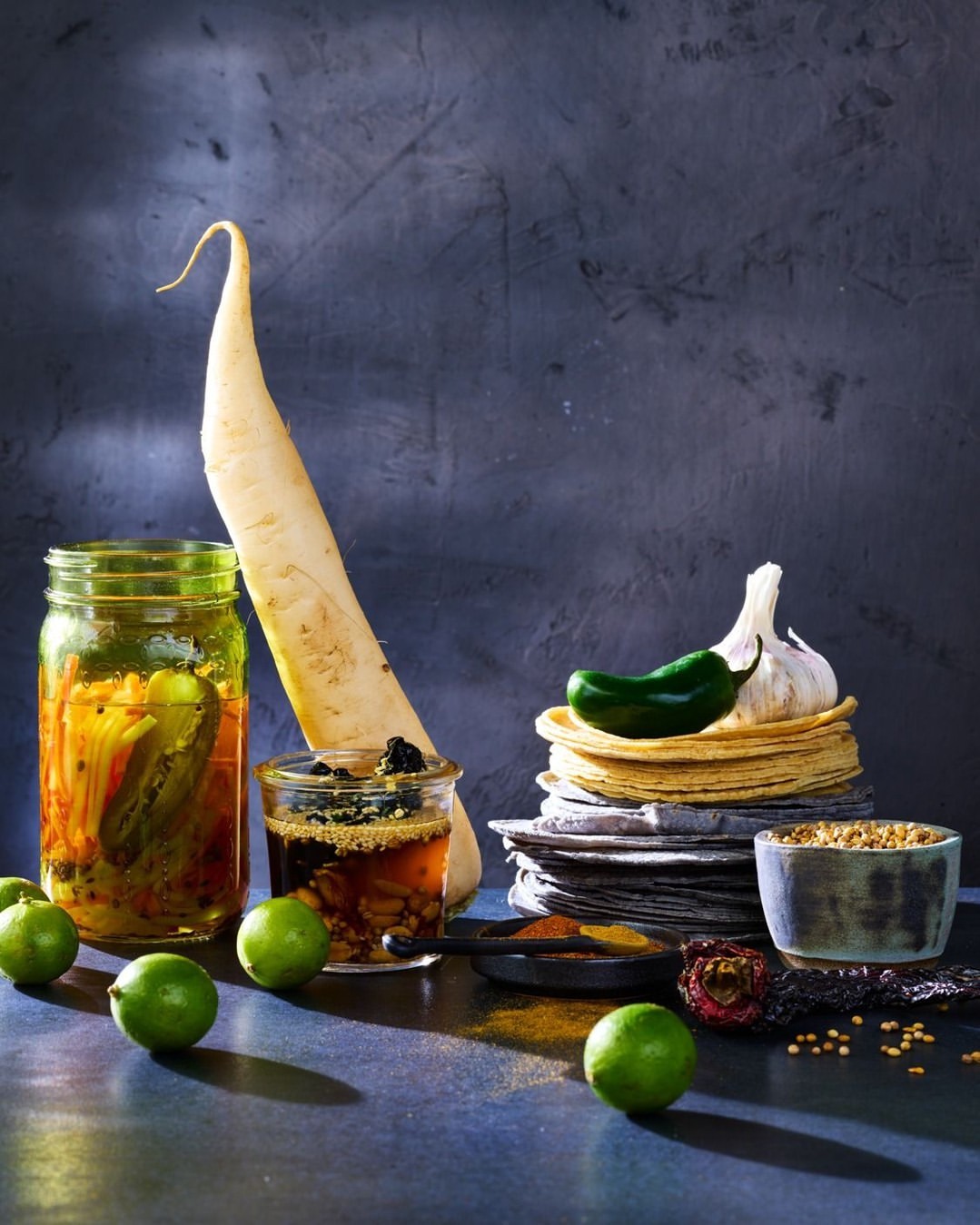 Still life food photography showcases pickled vegetables in glass jars, limes, tortillas, garlic, pepper, and spices against a dark background.