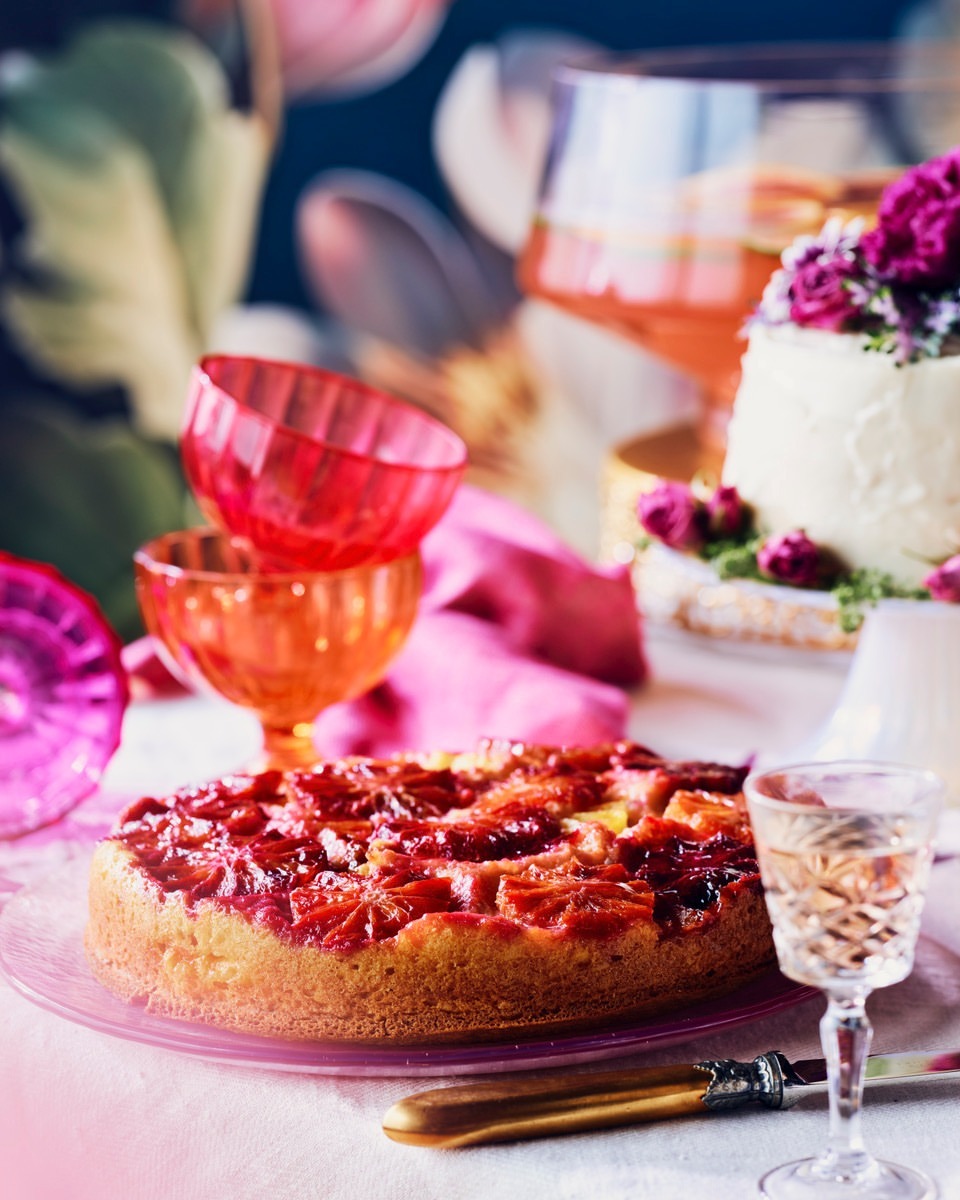 Overhead shot of a blood orange cake on a pink plate, surrounded by glassware and flowers on a white tablecloth, for a food editorial.