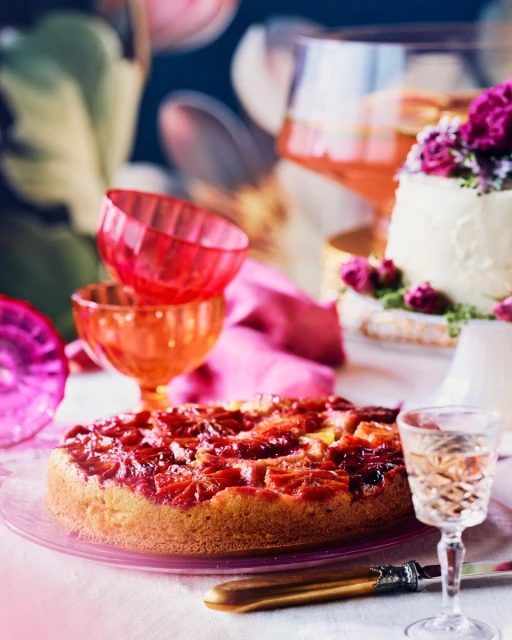 Overhead shot of a blood orange cake on a pink plate, surrounded by glassware and flowers on a white tablecloth, for a food editorial.