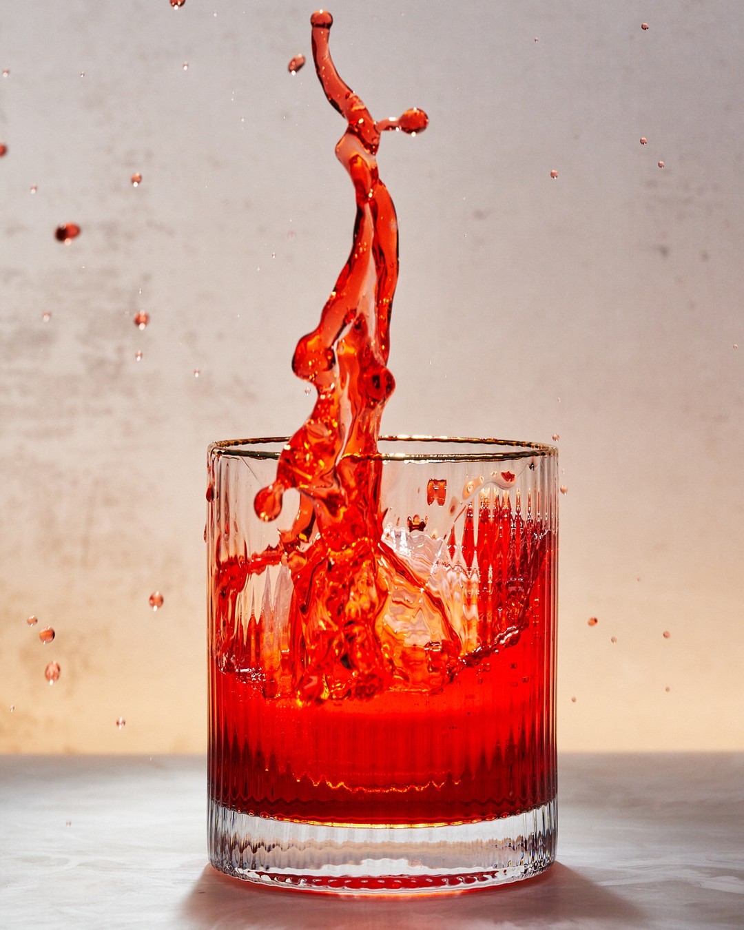 Close-up of a clear, ribbed glass filled with vibrant red liquid mid-splash, set against a neutral backdrop, highlighting its texture.