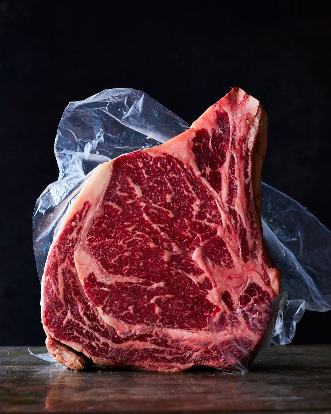 A large, raw cut of marbled beef ribeye steak encased in clear plastic film, resting on a dark wooden surface in a studio setting.
