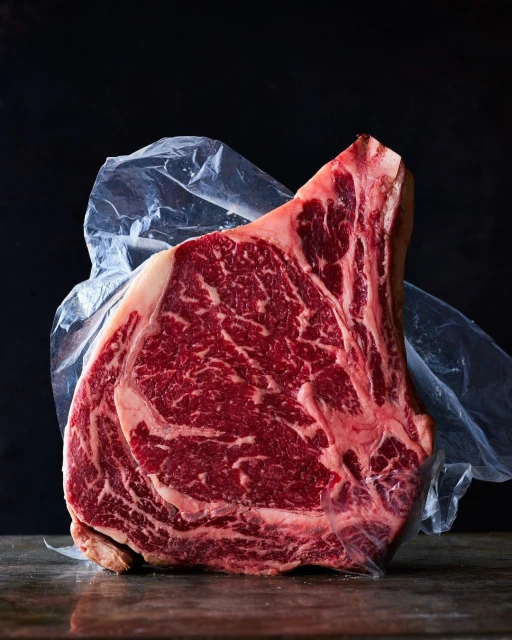 A large, raw cut of marbled beef ribeye steak encased in clear plastic film, resting on a dark wooden surface in a studio setting.