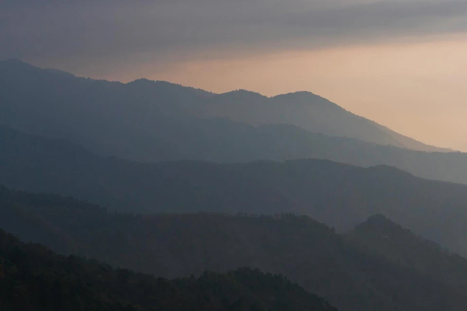 Scenic view of distant mountain range with dramatic cloudy sky, creating a hazy, atmospheric landscape at dusk or dawn.