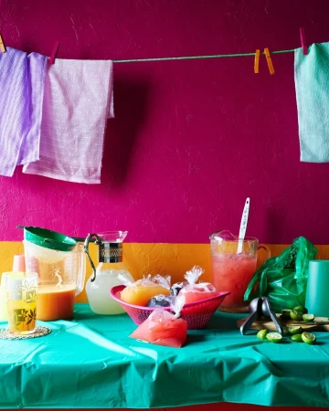 Colorful arrangement of fruit drinks in pitchers and plastic bags on a table with hanging towels, against a vibrant magenta wall.