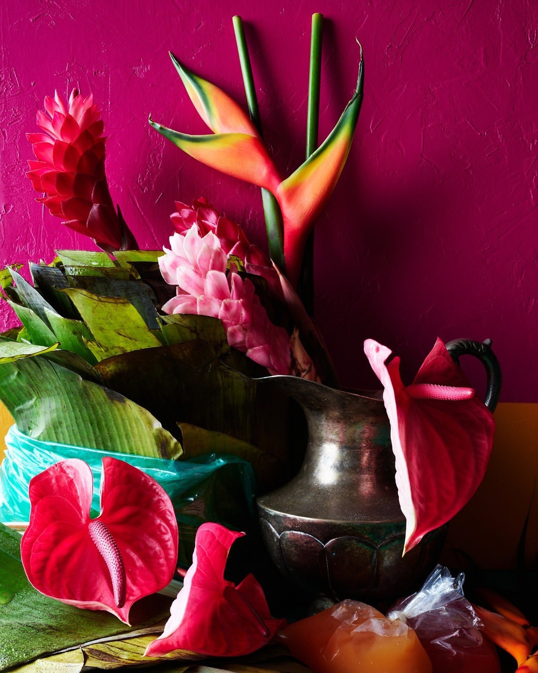 Still life with tropical flowers, including heliconia and anthurium, arranged in a silver pitcher against a magenta background.
