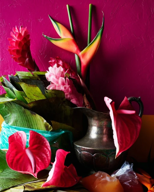 Still life with tropical flowers, including heliconia and anthurium, arranged in a silver pitcher against a magenta background.